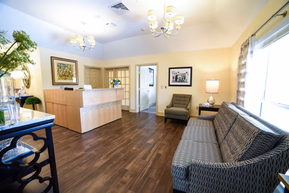 A bright and welcoming reception area in a senior living facility featuring a wooden reception desk, a patterned sofa, an armchair, a side table with a lamp, framed artwork on the walls, and large windows with curtains allowing natural light to fill the room.