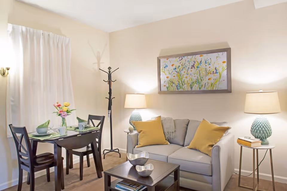 A cozy living room and dining area with a small dining table set for two, a gray loveseat with yellow pillows, two table lamps with textured bases, a coat rack, and a colorful floral painting on the wall.