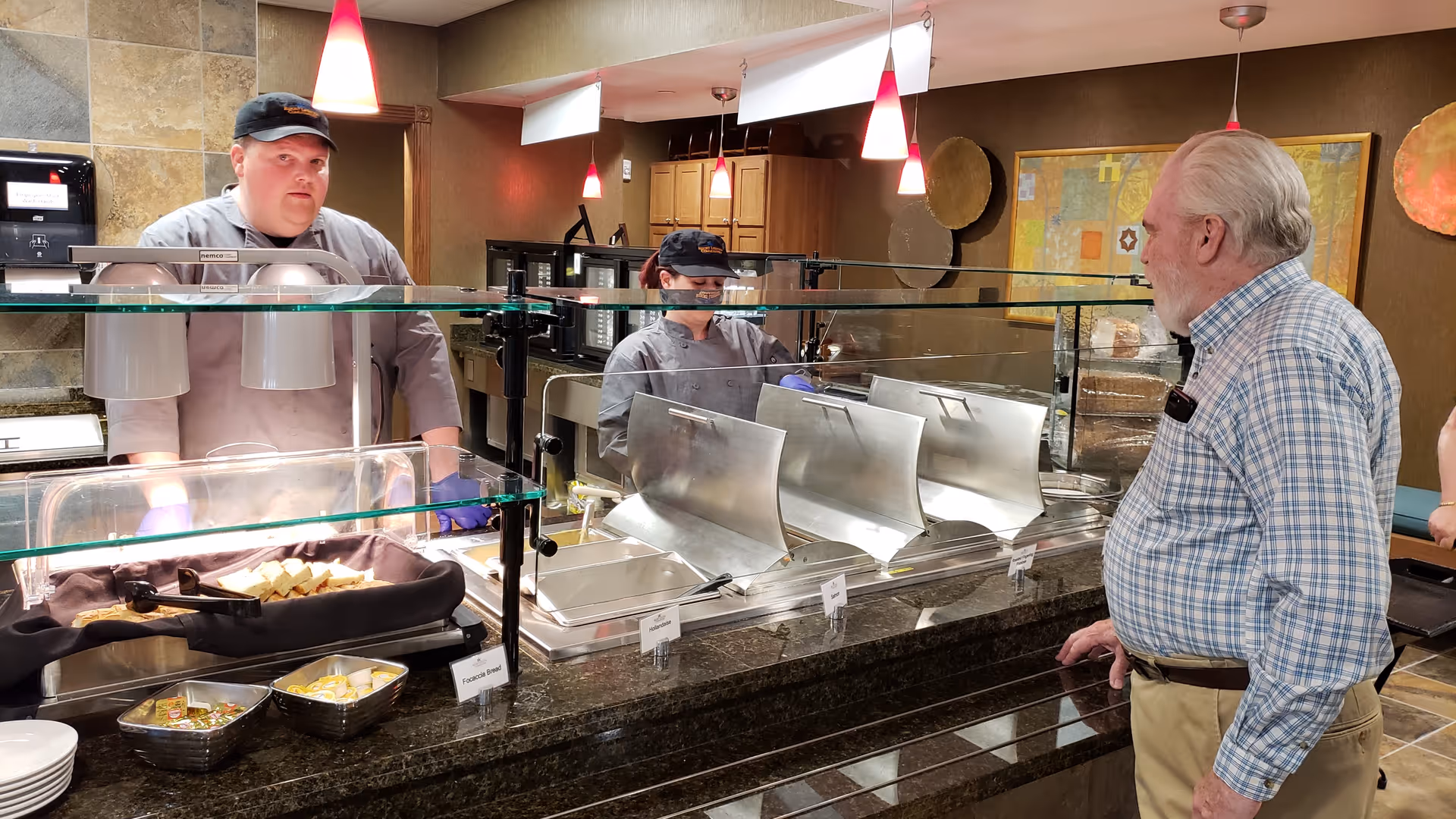 An elderly man stands at a cafeteria-style serving counter while two staff members behind a glass sneeze guard prepare and serve food.