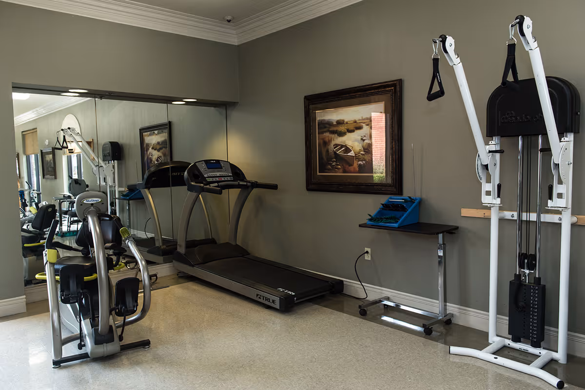 A small fitness room with exercise equipment including a treadmill, a recumbent bike, and a weight machine. The room has a large mirror on one wall, a framed painting of a boat on a lake, and a small table with exercise accessories. The walls are painted gray and the floor is light-colored.