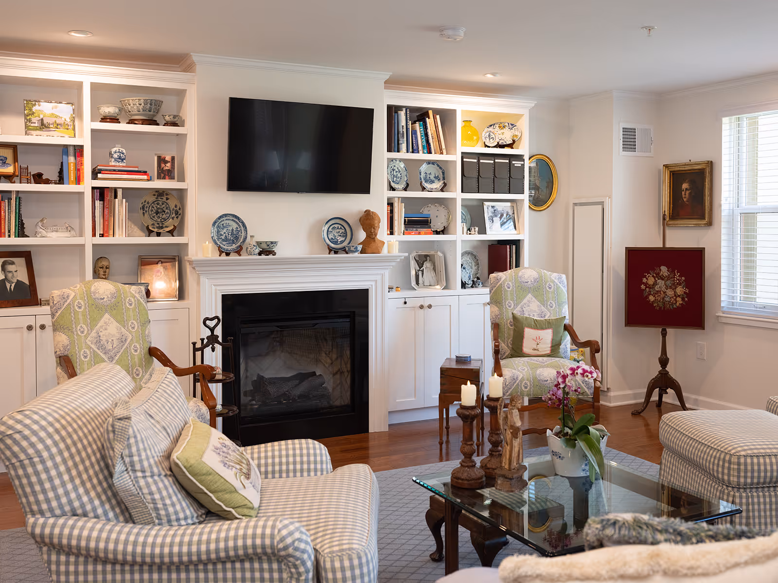 A cozy living room with patterned armchairs and a glass coffee table with decorative items and a potted orchid. The room features a fireplace with a mounted flat-screen TV above it, flanked by built-in white shelves filled with books, plates, and decorative objects. There are framed pictures and paintings on the walls, and windows letting in natural light.