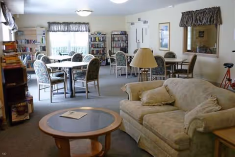 Cozy senior living common room with beige sofa, round coffee table, and group dining tables.