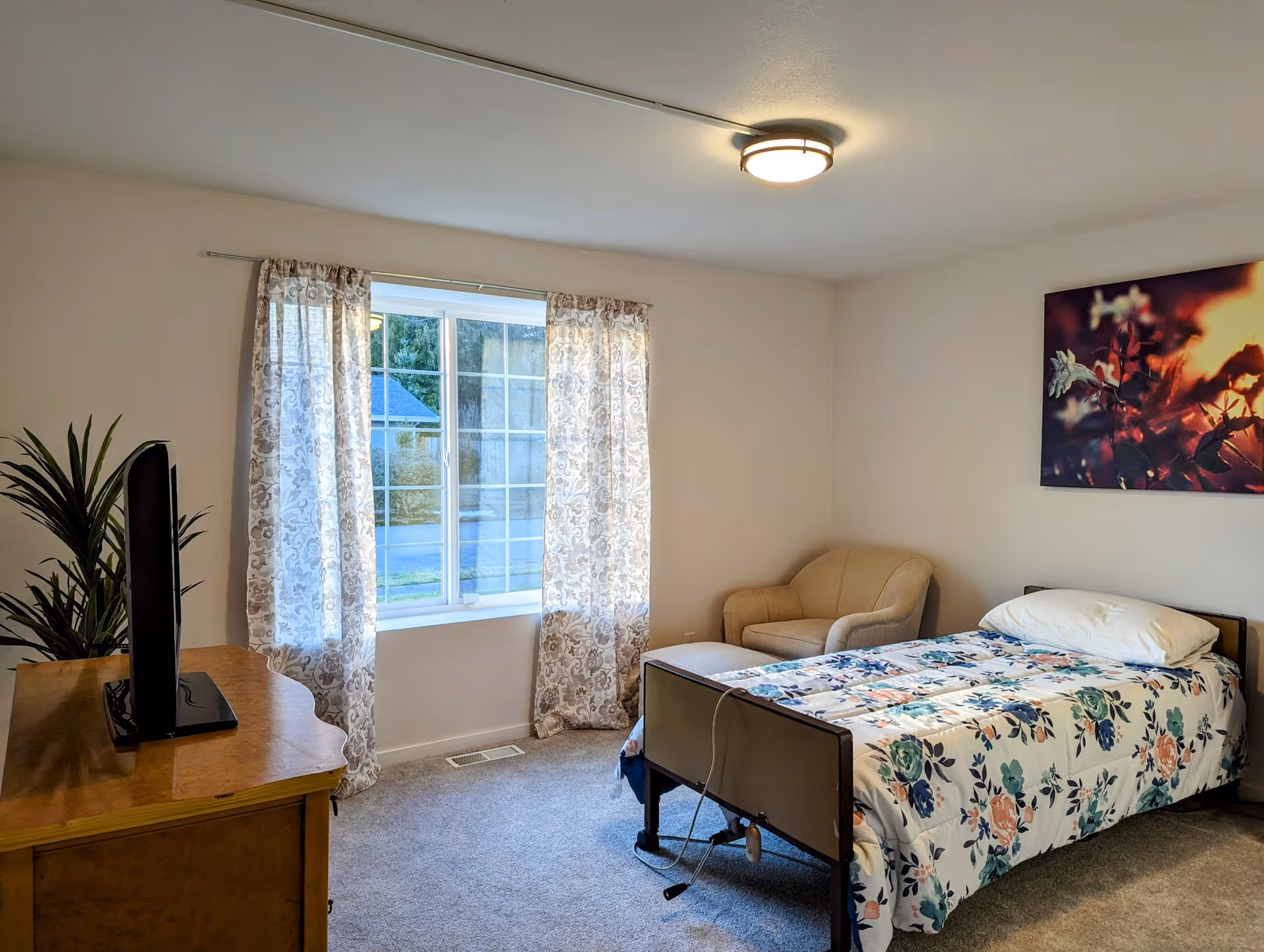 A bedroom with a single bed covered in a floral patterned comforter, a beige armchair in the corner, a wooden dresser with a flat-screen TV on top, a large window with patterned curtains, and a colorful floral artwork on the wall.