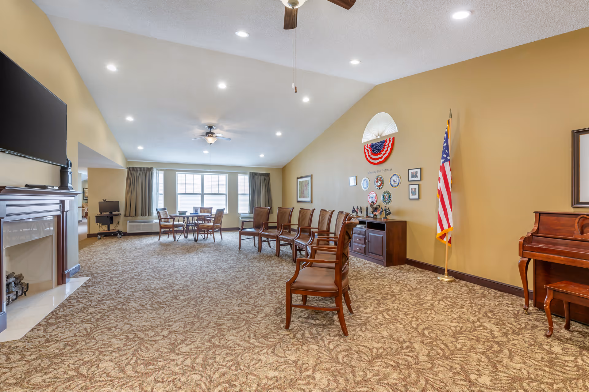 A spacious common room with beige patterned carpet and tan walls. There are several wooden chairs with cushions arranged in a row facing a fireplace with a mounted TV above it. At the far end of the room, there is a round table with four chairs near large windows with curtains. On the right side, there is a wooden piano and an American flag standing next to a cabinet decorated with patriotic items and wall plaques. The ceiling has recessed lighting and ceiling fans.
