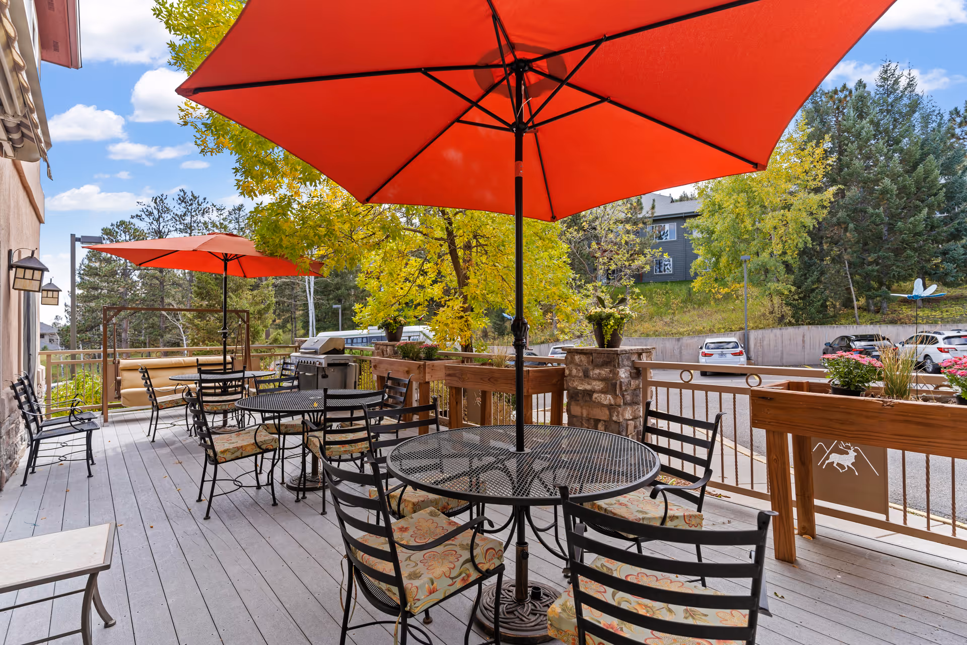 Outdoor patio area with several black metal tables and chairs with floral cushions, shaded by large red umbrellas. The patio overlooks a parking lot with cars and is surrounded by trees with green and yellow leaves under a partly cloudy sky.