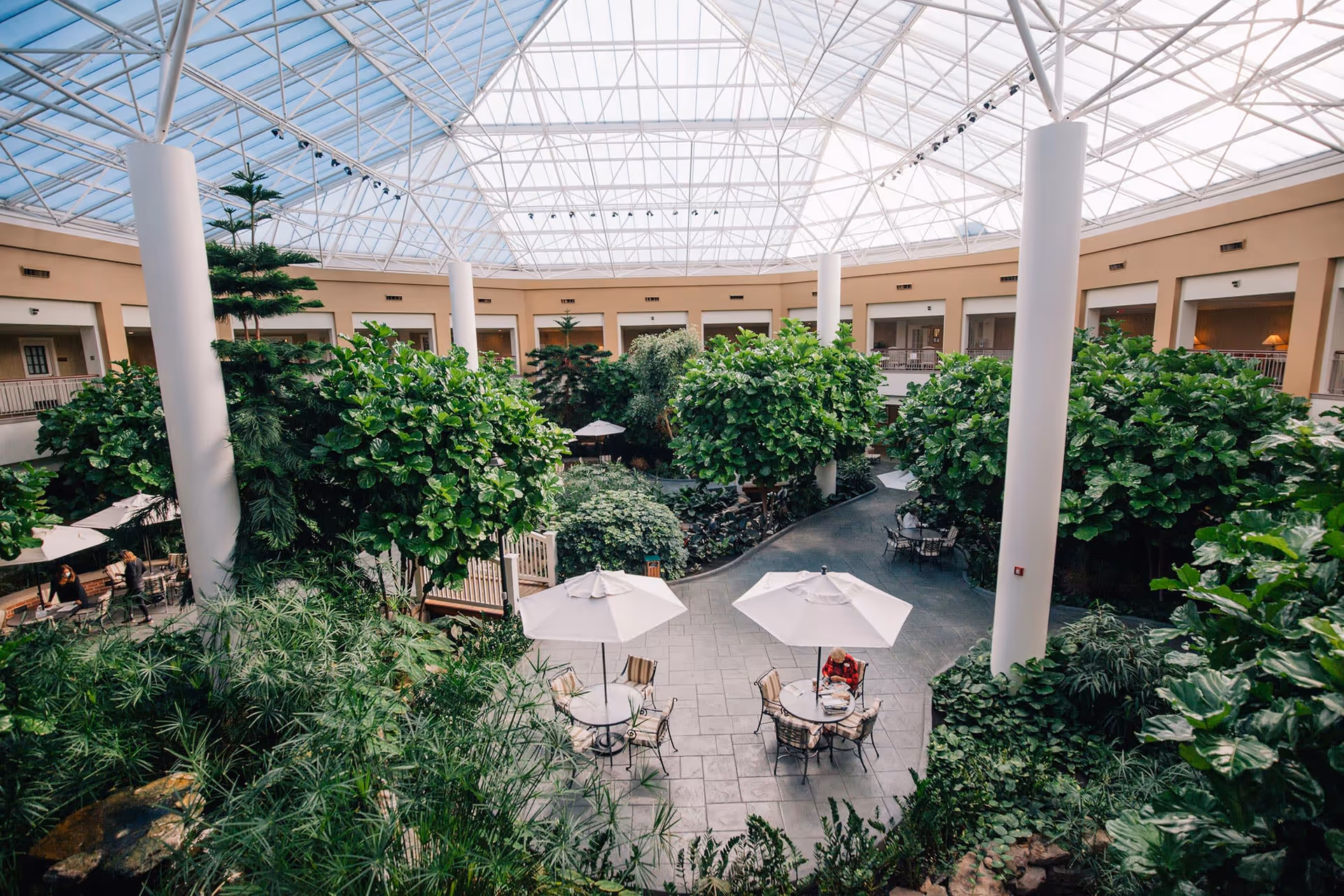 Indoor atrium with lush green plants and trees under a large glass ceiling. There are several round tables with umbrellas and chairs arranged on a tiled floor. A few people are seated and walking around the area. The atrium is surrounded by a two-story building with balconies overlooking the space.