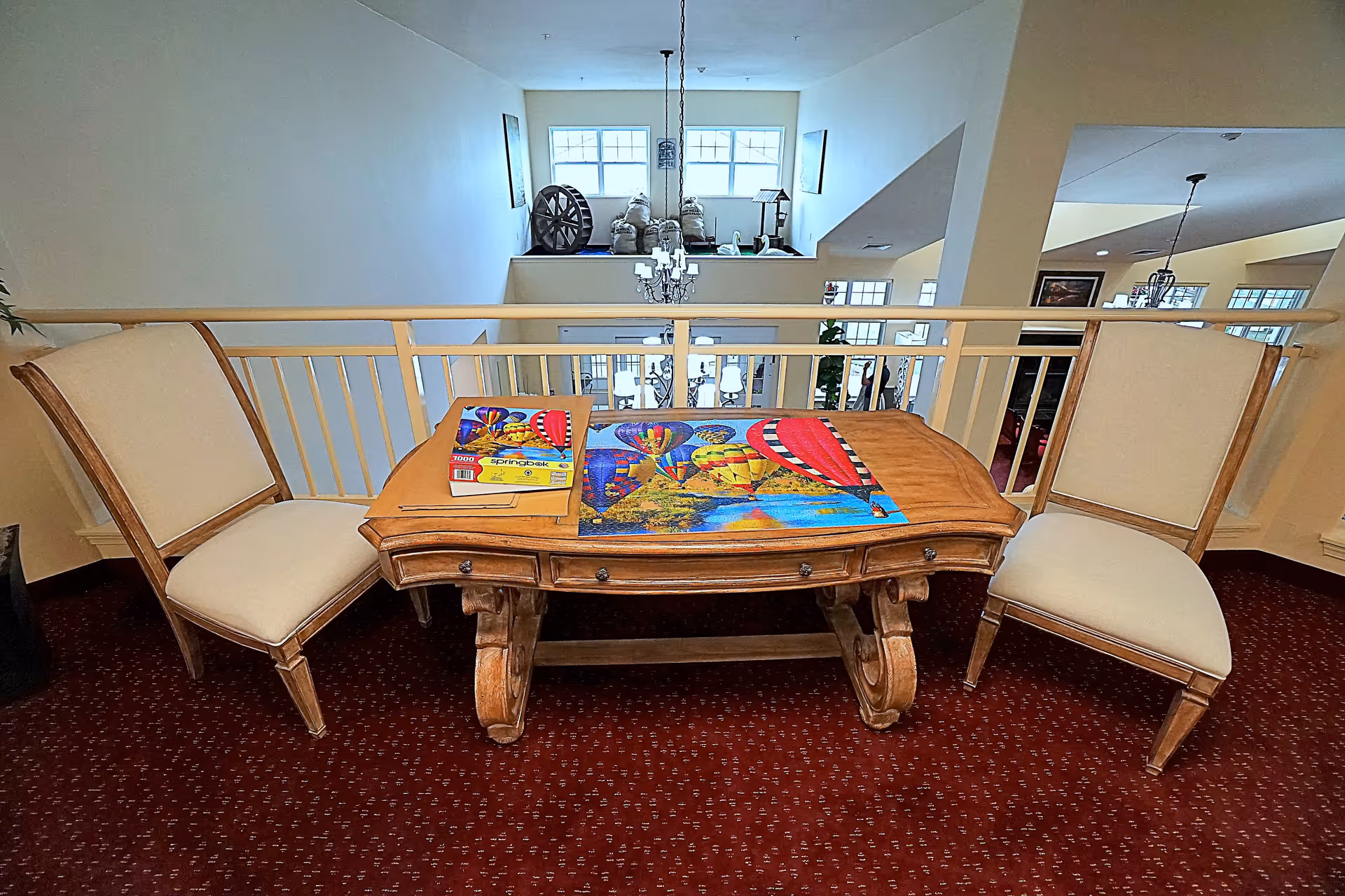 A wooden table with a colorful hot air balloon puzzle partially completed on top, flanked by two beige cushioned chairs. The setting is an upper-level interior space with a railing overlooking a lower floor with large windows and a chandelier.