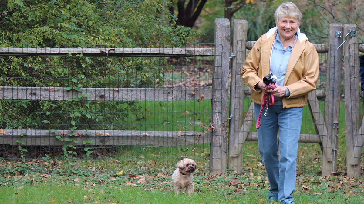 An elderly woman smiling while walking a small dog beside a wooden fence in a grassy outdoor area.
