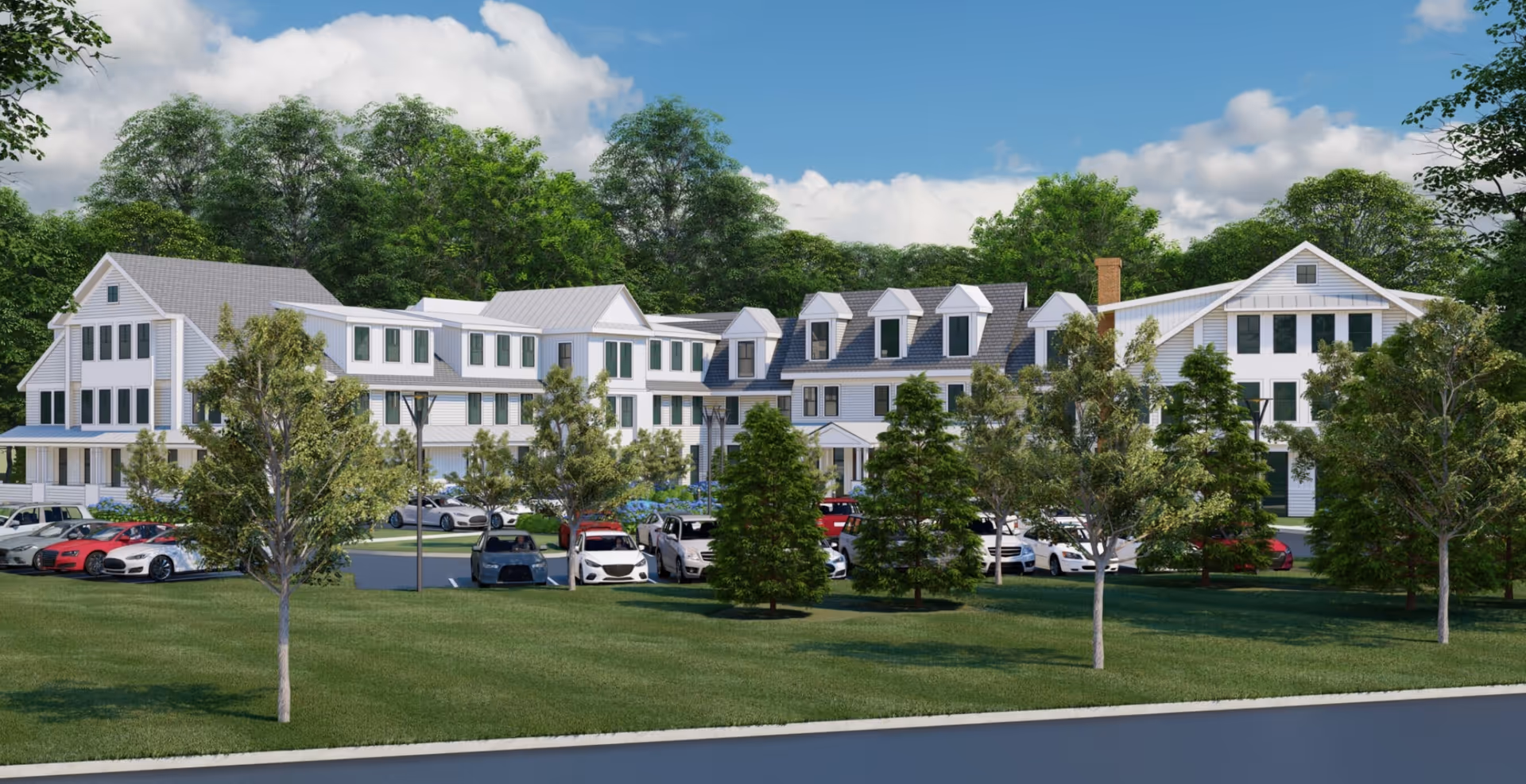 Exterior view of a large, multi-story senior living facility building surrounded by trees and a parking lot with several cars parked. The building has white siding, multiple windows, and a pitched roof under a partly cloudy blue sky.