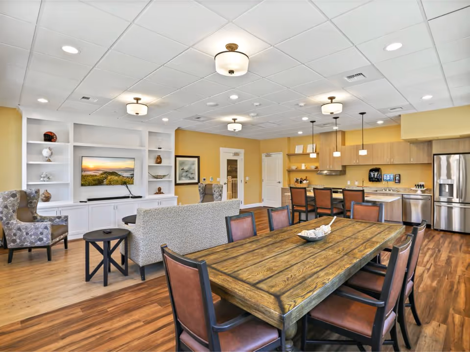 Open communal dining and living area with a long wooden dining table, chairs, a seating area with sofa and armchairs, and a kitchenette in the background.