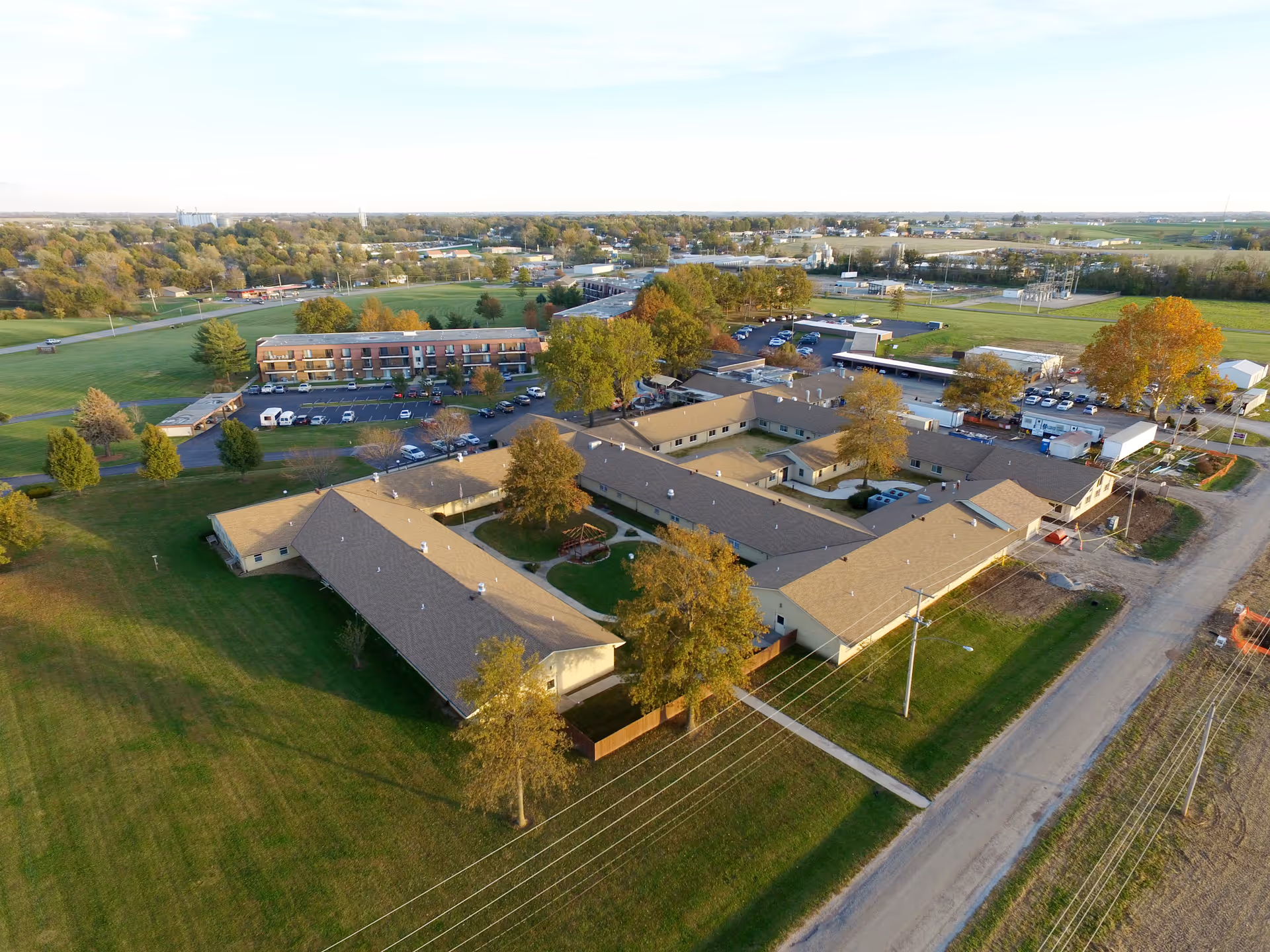 Aerial view of John Knox Village East senior living facility showing multiple connected buildings with beige roofs surrounded by green lawns, trees, parking lots, and nearby roads under a clear sky.