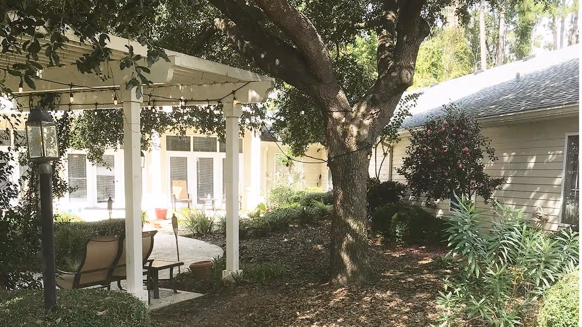 Outdoor garden area at Village Cove Assisted Living featuring a large tree, a white pergola with string lights, patio chairs, and surrounding greenery including bushes and flowering plants, adjacent to a light-colored building.