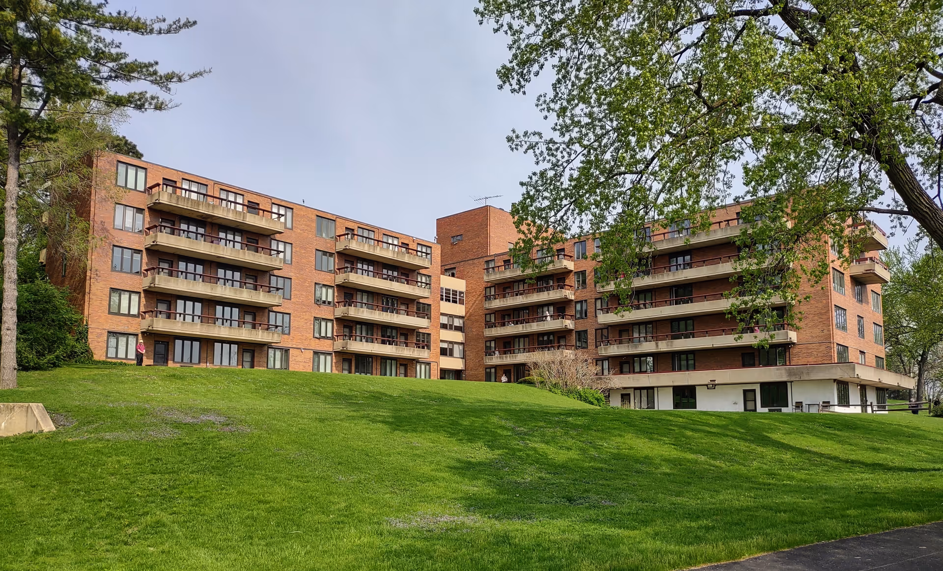 Exterior view of a multi-story brick building with balconies, surrounded by green grass and trees under a clear sky.