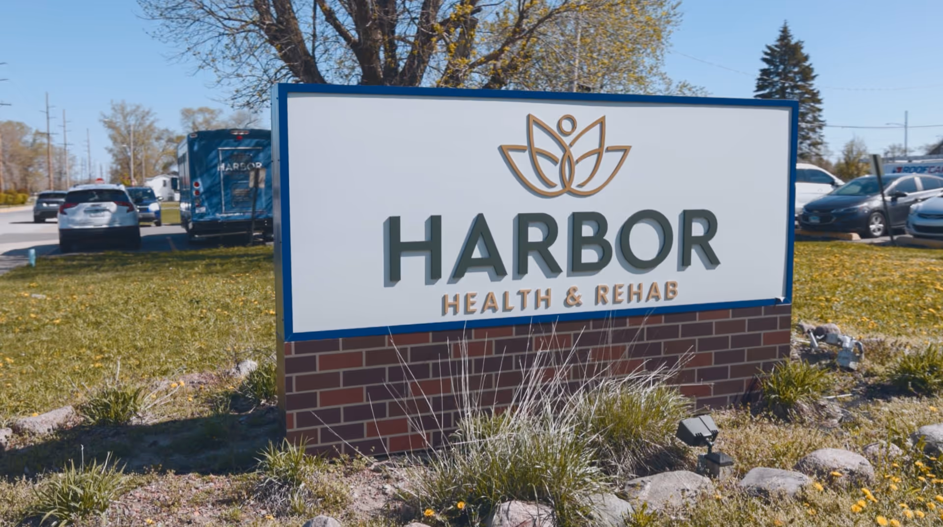Outdoor view of a sign for Harbor Health & Rehab, featuring a stylized lotus flower logo above the facility name. The sign is mounted on a brick base and surrounded by grass and small plants, with parked cars and trees visible in the background.