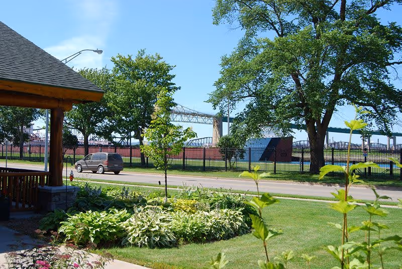 View of a landscaped garden area with green plants and trees, a road with a parked vehicle, and a large bridge structure in the background under a clear blue sky. Part of a wooden building structure is visible on the left side.
