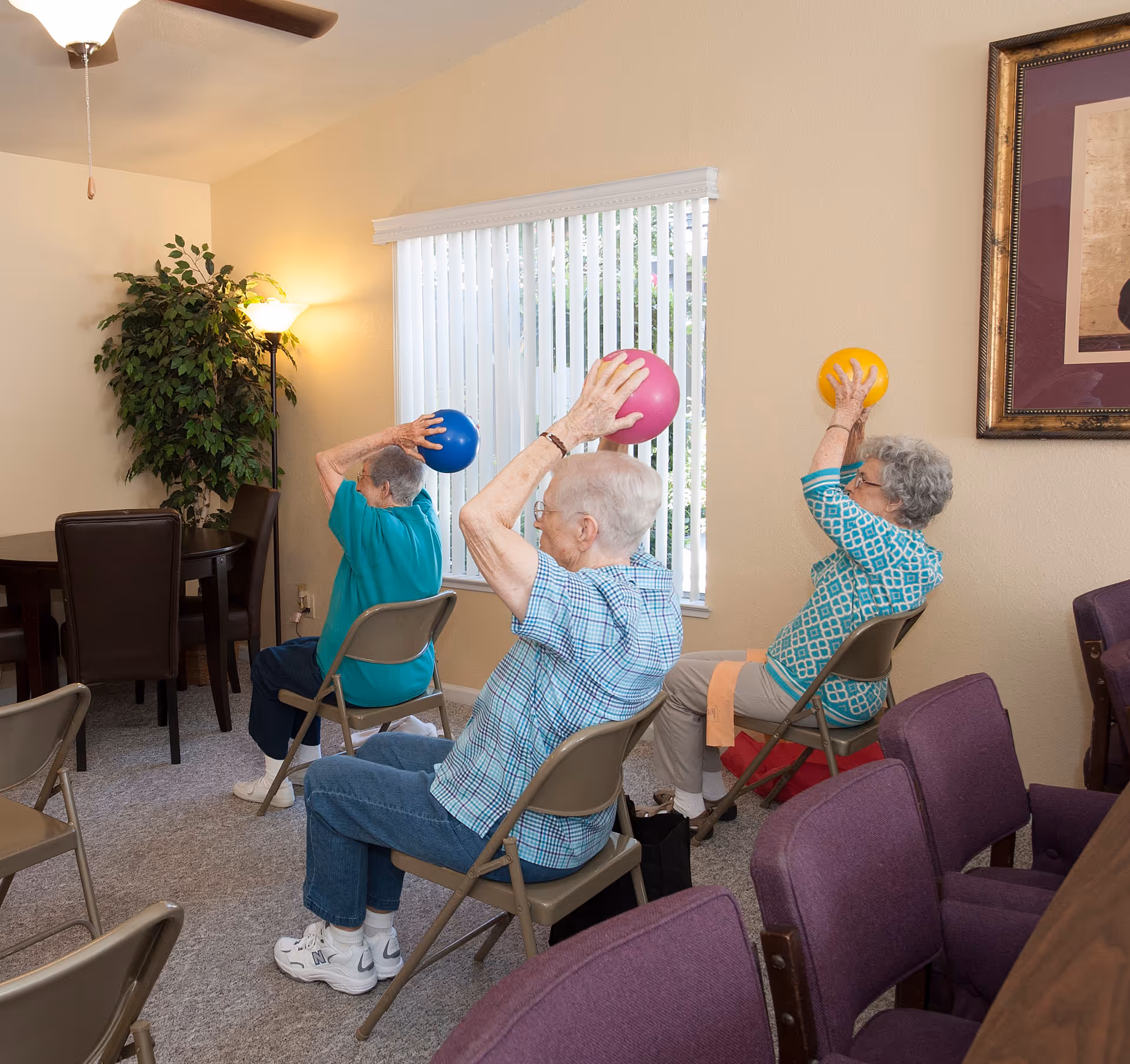Three elderly women seated on folding chairs in a room, each holding a small exercise ball above their heads as part of a seated exercise activity. The room has beige walls, a window with vertical blinds, a ceiling fan, a floor lamp, a potted plant, and framed artwork on the wall.