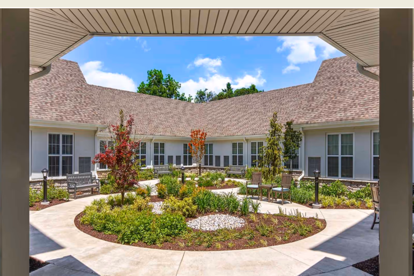 A landscaped courtyard garden with a circular walkway, benches, chairs, small trees, and various plants, surrounded by a building with multiple windows under a clear blue sky.