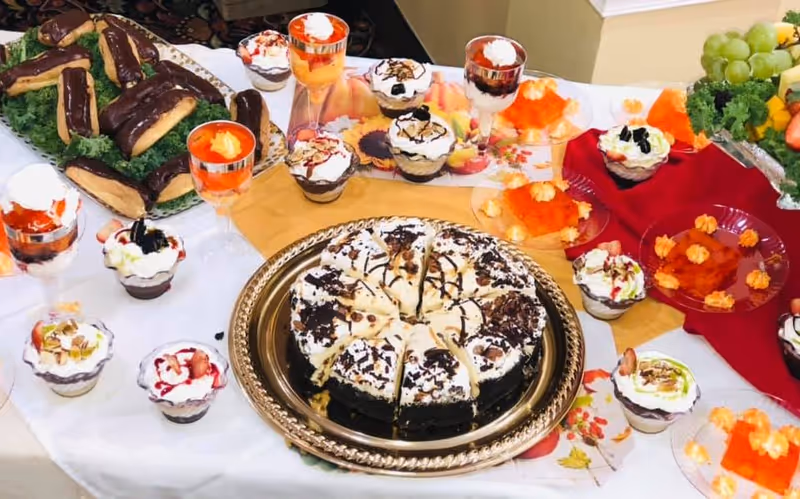 A table set with an assortment of desserts including a sliced chocolate cake with white frosting and chocolate drizzle on a silver platter, several small dessert cups with whipped cream and fruit toppings, éclairs covered in chocolate on a bed of greens, and plates with orange gelatin topped with whipped cream. There is also a fruit arrangement with grapes and strawberries on the side.