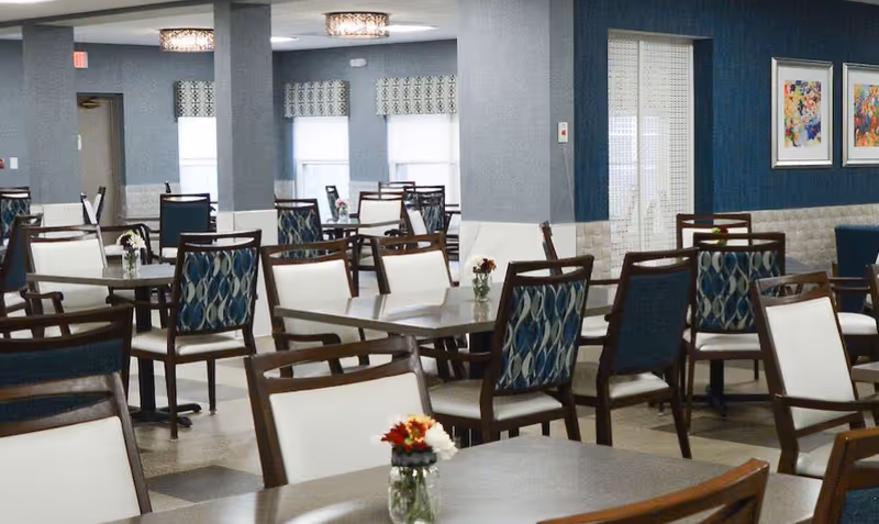 A spacious dining room with multiple tables and chairs arranged neatly. The chairs have wooden frames with white and patterned blue upholstery. Small flower vases are placed on the tables. The room has blue and white walls, windows with patterned valances, and framed artwork on the walls.