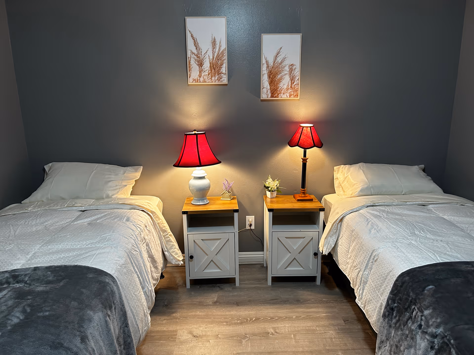 Two twin beds separated by matching nightstands with lit table lamps against a gray wall with framed artwork.