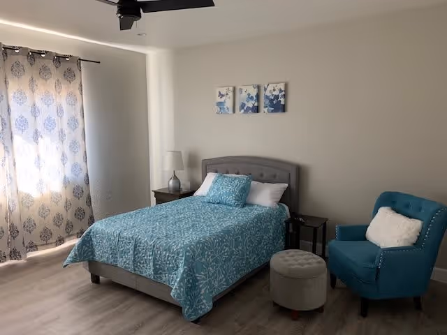 A bright bedroom with a double bed dressed in teal bedding, a nightstand and lamp, a teal armchair, ottoman, and patterned curtains.