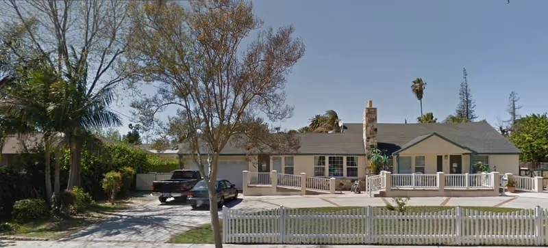 Front exterior view of a single-story residential building with a white picket fence, a driveway with two parked vehicles, and several trees and shrubs in the yard under a clear blue sky.