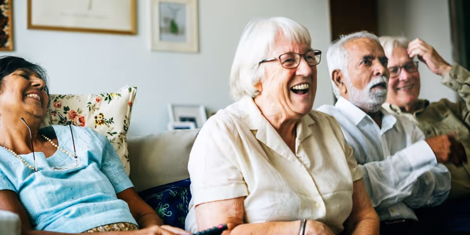Four elderly people sitting on a couch in a living room, smiling and laughing together. The group includes two women and two men, with a cozy and friendly atmosphere.