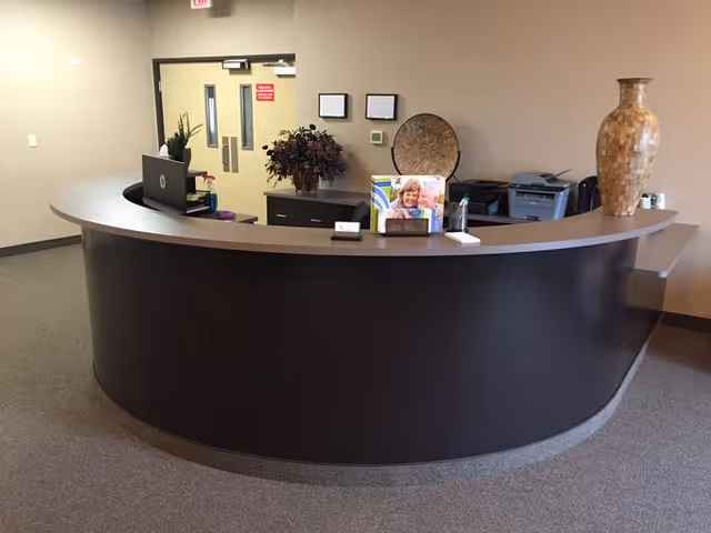 A curved reception desk in a lobby with office doors, decorative vase, printer, plants and a brochure on the counter.