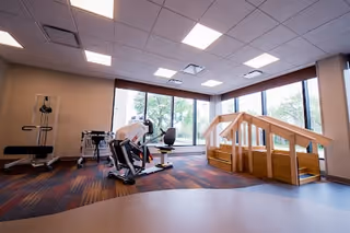 A senior living community exercise room with large windows letting in natural light. The room contains exercise equipment including a stationary bike and a set of wooden stairs with handrails for physical therapy or rehabilitation exercises. The floor has a mix of carpet and smooth surfaces, and the ceiling has recessed lighting.