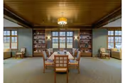 A spacious, well-lit common area with a wooden ceiling and carpeted floor. The room features a central table surrounded by chairs, flanked by built-in bookshelves filled with books and decorative items. Large windows on the far wall allow natural light to enter, and additional seating is placed near the windows on either side.