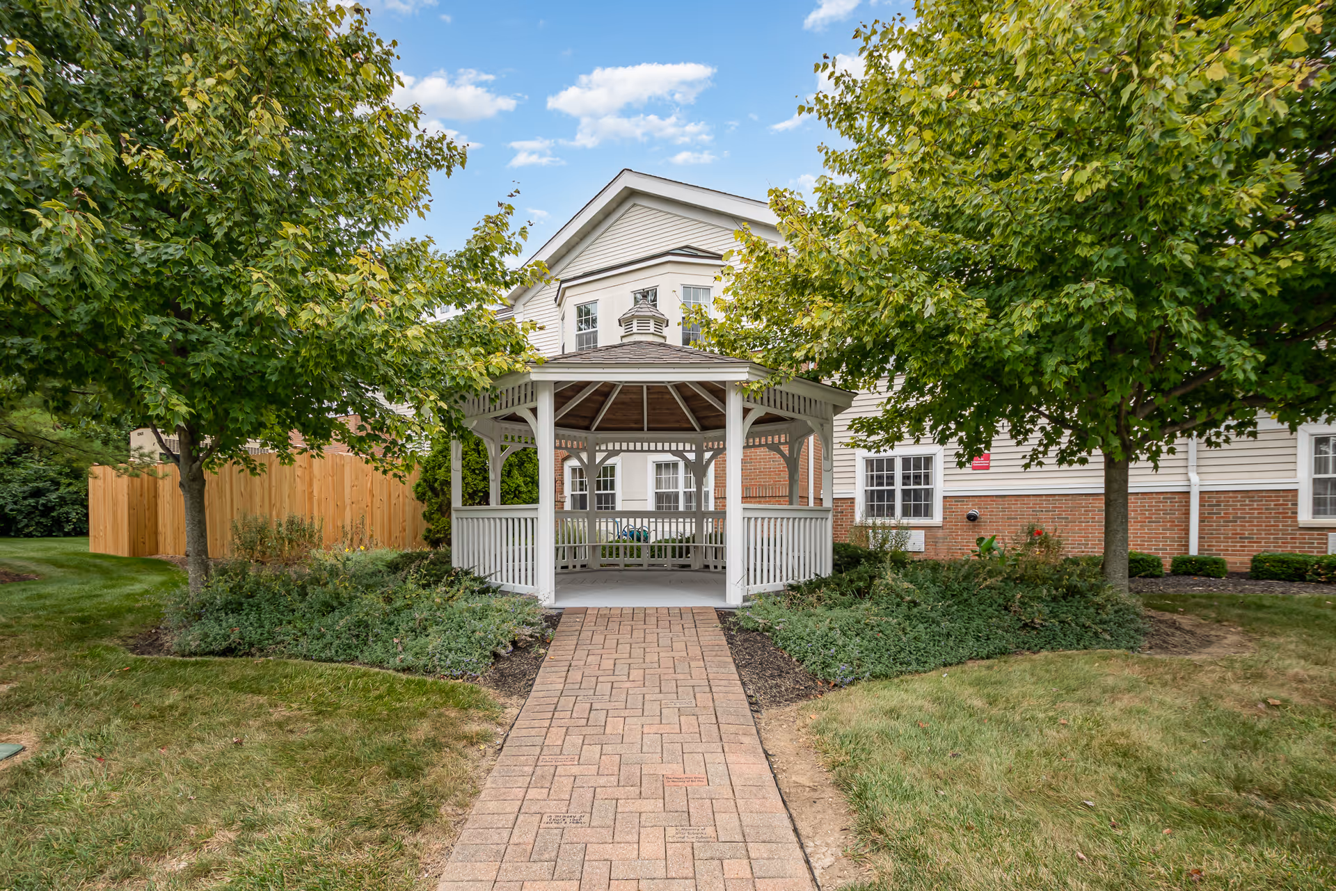 A white wooden gazebo with a shingled roof is centered at the end of a brick pathway, surrounded by green grass, bushes, and trees. Behind the gazebo is a two-story building with white siding and red brick on the lower half. The sky is blue with some clouds.