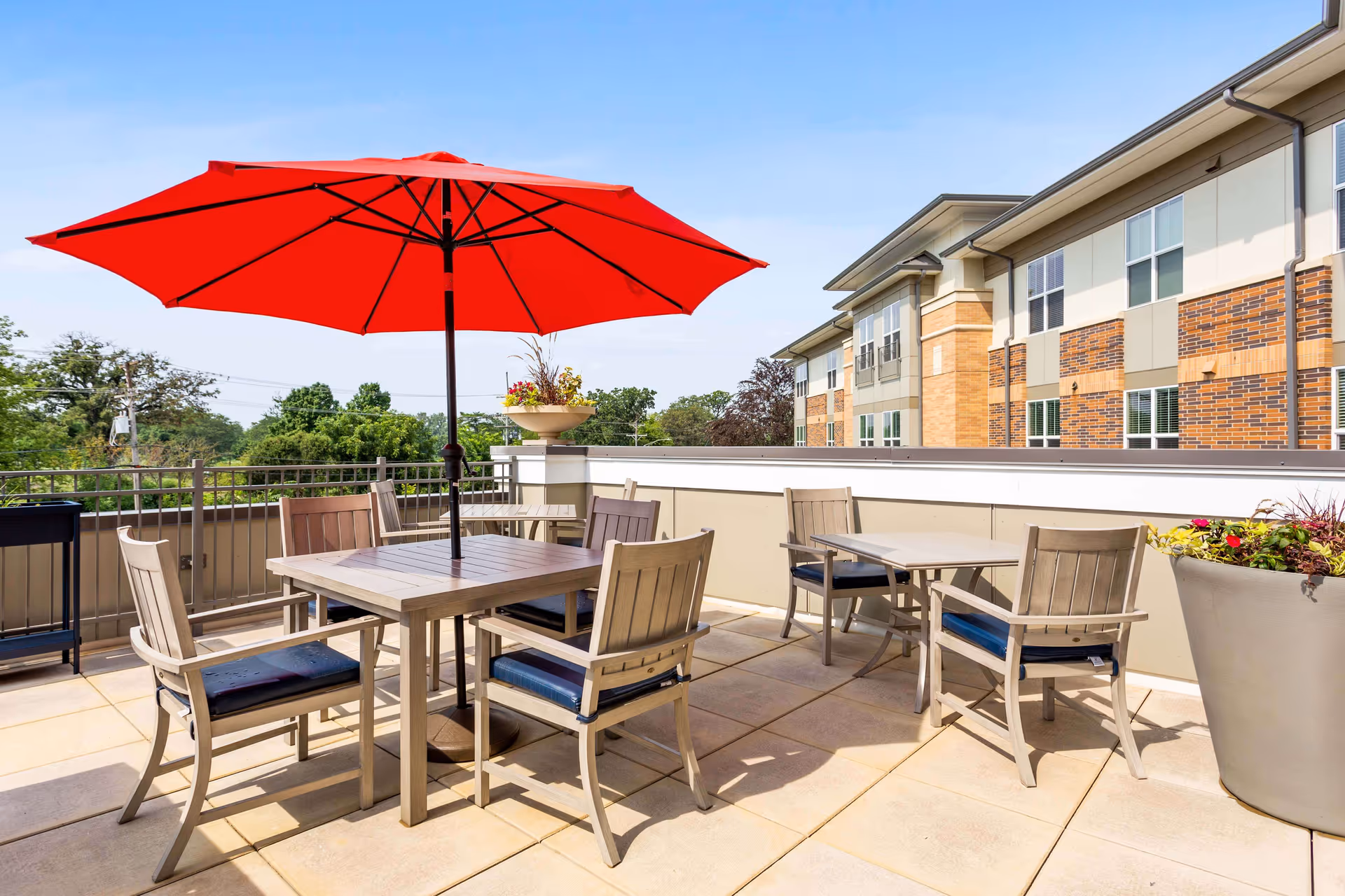 Outdoor patio area with several tables and chairs, one table featuring a large red umbrella. The patio is surrounded by a railing and overlooks greenery and part of a multi-story building with brick and beige siding.