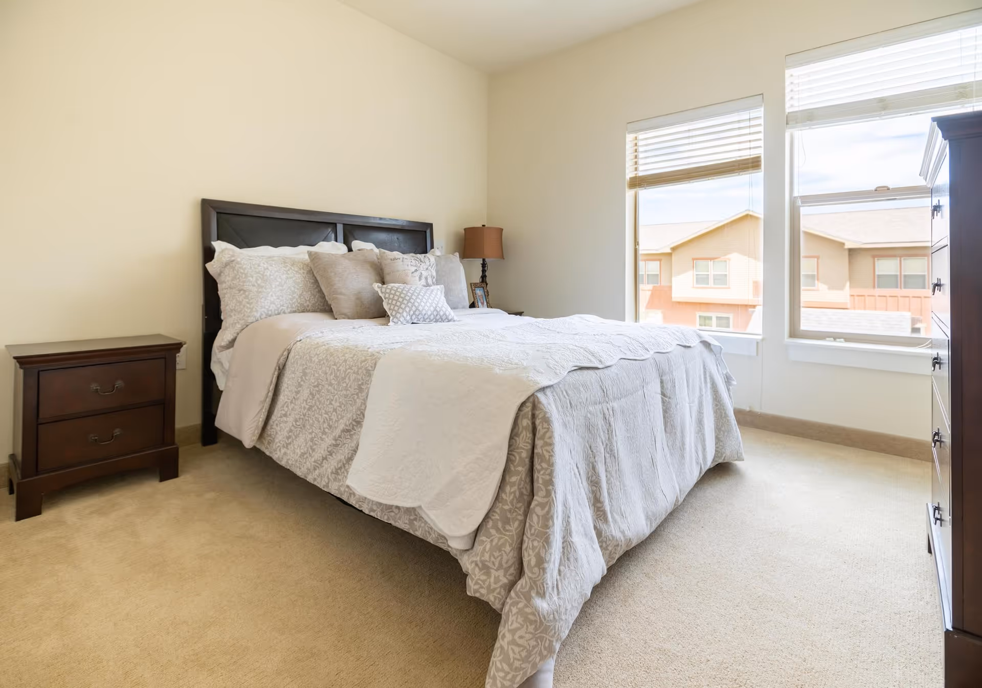Sunlit bedroom with a neatly made bed, nightstand, lamp and dresser beside two large windows.
