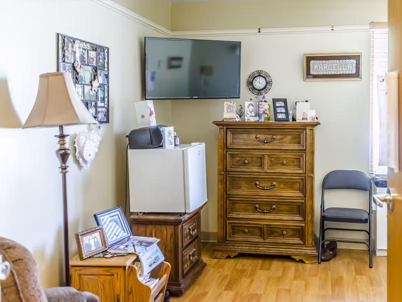 A cozy room with wooden furniture including a tall dresser and a small cabinet holding a mini fridge and a radio. A flat-screen TV is mounted on the wall above the fridge. There is a floor lamp on the left side and a folding chair next to the dresser on the right. The walls are decorated with framed photos, a clock, and a framed name plaque that reads 'Marcella'. The floor is wooden, and the room has a warm, homey feel.