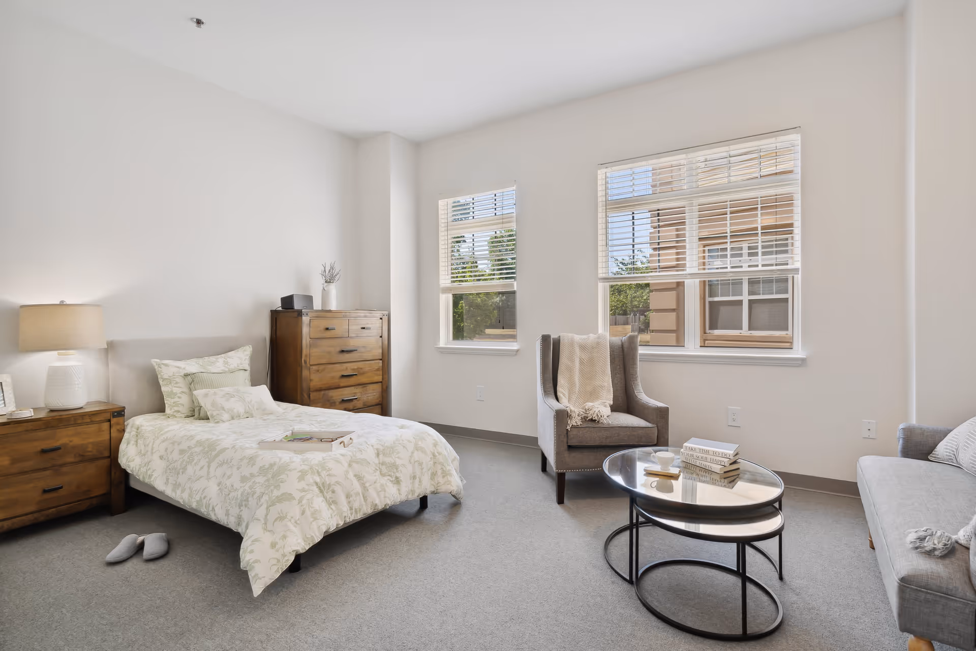 A bright and cozy bedroom in an assisted living facility featuring a single bed with floral bedding, a wooden nightstand with a lamp, a wooden dresser, a comfortable armchair with a throw blanket, a round glass coffee table with books and a cup, and a gray sofa. Two windows with white blinds allow natural light to fill the room.