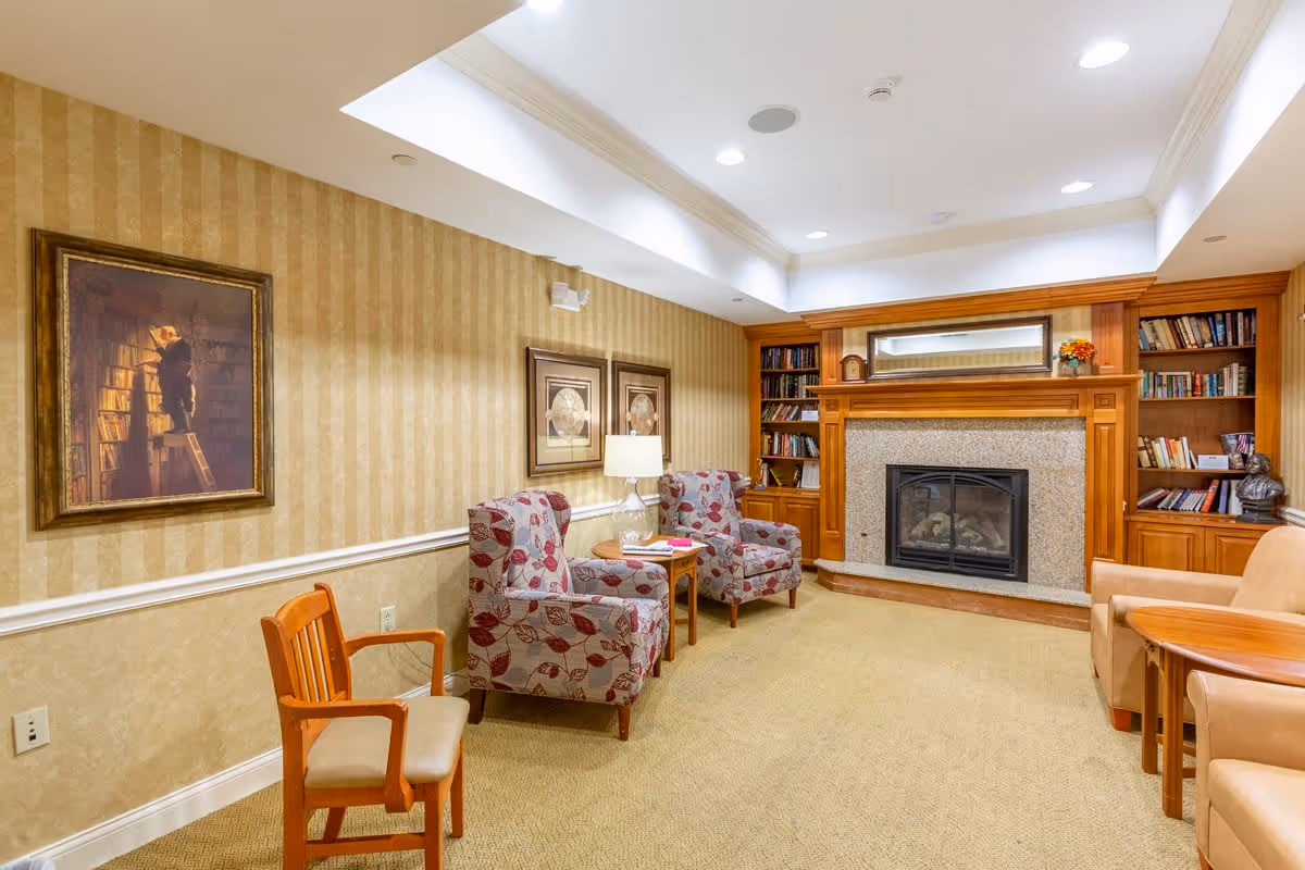 A cozy living room area with patterned armchairs, a wooden chair, a small round table with a lamp, and a fireplace surrounded by wooden built-in bookshelves filled with books and decorative items. The walls have striped wallpaper and framed artwork, and the ceiling has recessed lighting.