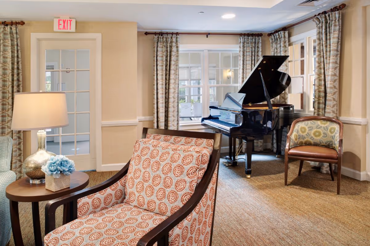 Cozy senior living common room with patterned armchairs, a side table lamp, and a grand piano by the windows.
