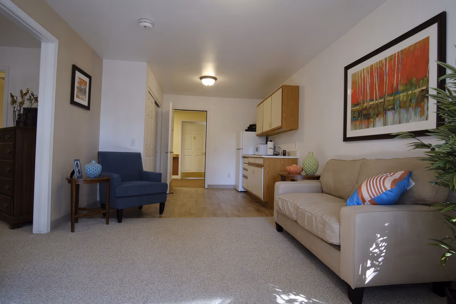 A cozy living area in a senior living facility featuring a beige sofa with a colorful pillow, a blue armchair next to a small wooden side table with a blue vase and a framed photo. The room has light-colored walls with framed artwork, a small kitchenette with wooden cabinets, a refrigerator, microwave, and sink. The floor transitions from carpet in the living area to wood in the kitchenette. A door is open leading to another room.