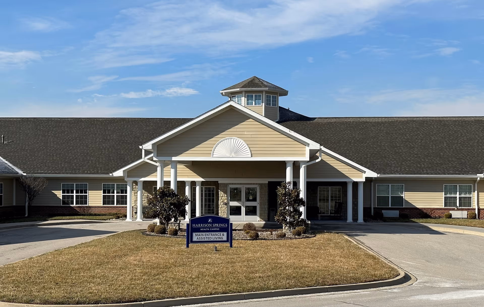 Front exterior view of Harrison Springs Health Campus building with a covered entrance supported by white columns, a small landscaped area with shrubs, and a sign indicating main entrance and assisted living. The sky is clear with some clouds.
