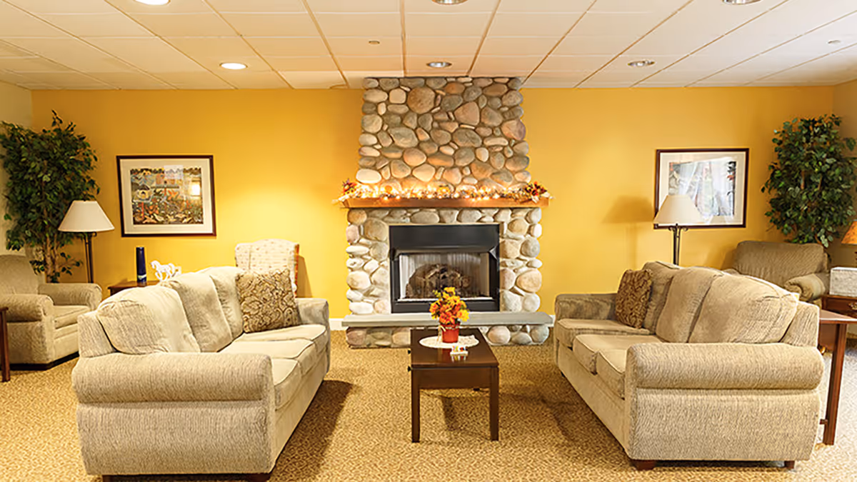 A cozy living room with two beige sofas facing each other across a wooden coffee table with a small flower arrangement. The room features a stone fireplace with a wooden mantle decorated with string lights and small autumn decorations. The walls are painted yellow and adorned with framed artwork. There are two floor lamps and potted plants in the corners, creating a warm and inviting atmosphere.
