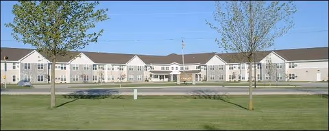 Long two-story assisted living building facade with a wide lawn, two trees in the foreground, and a clear blue sky.