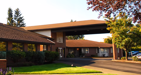 Exterior view of a single-story senior living facility building with a covered entrance, surrounded by green bushes, trees, and a well-maintained lawn under a clear sky.