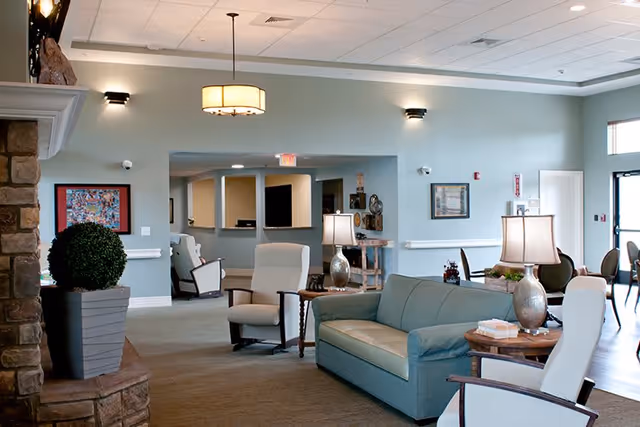 A spacious and well-lit senior living facility common area with comfortable seating including a teal sofa and beige armchairs. There are side tables with lamps, a large potted plant, and framed artwork on the light blue walls. The room has a stone fireplace on the left and a dining area visible in the background near large windows.
