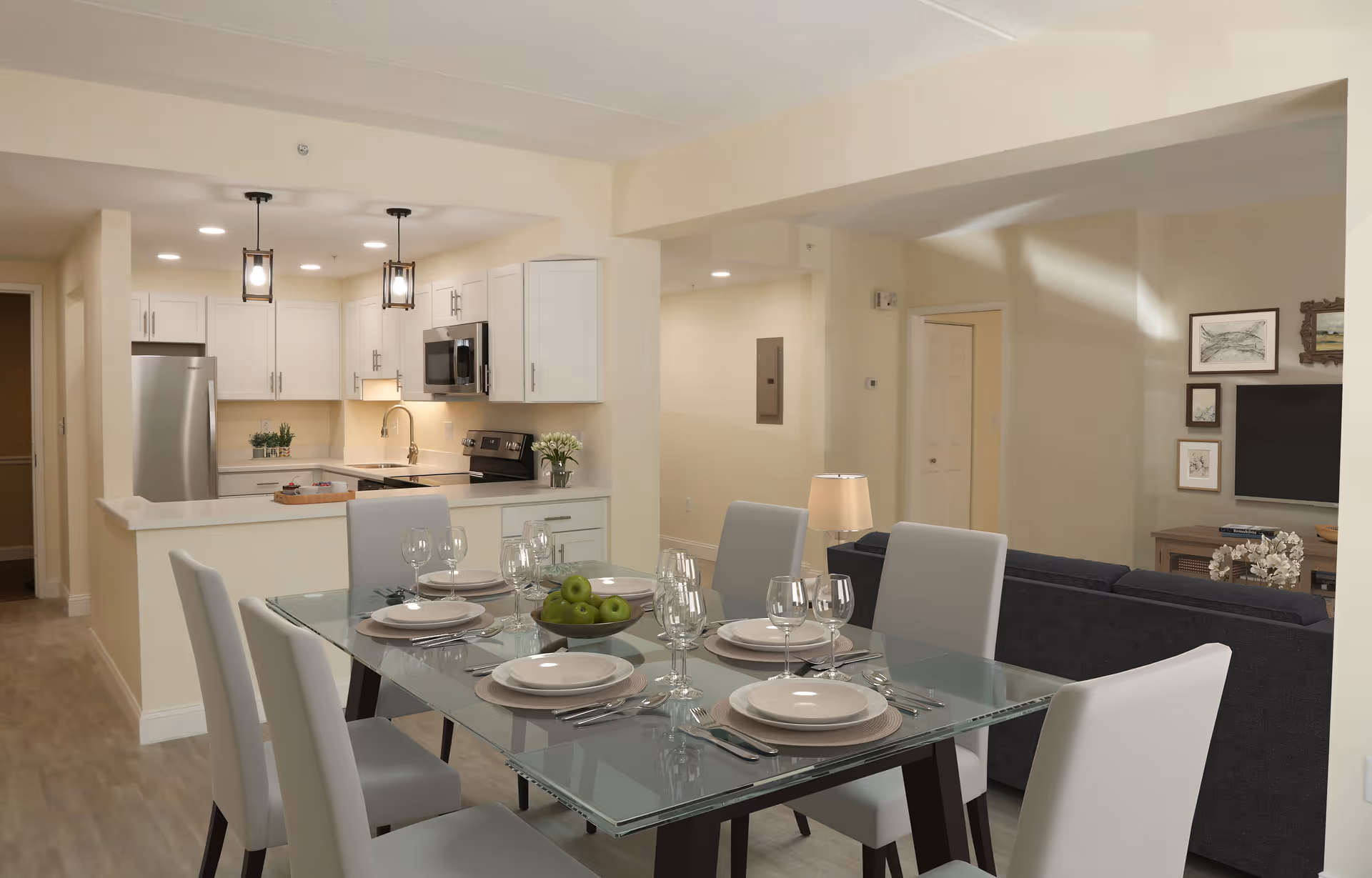 Modern open-concept dining and kitchen area with a glass dining table set for six, white chairs, and a kitchen featuring white cabinets, stainless steel appliances, and pendant lights.