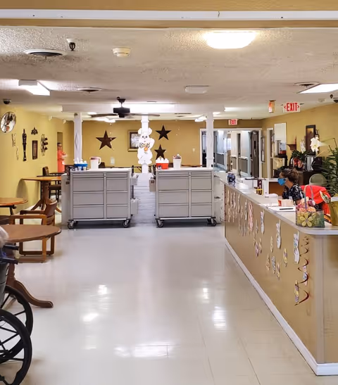 Interior view of a nursing home hallway with a reception desk on the right decorated with paper cutouts. Two large mobile carts are positioned in the center of the hallway. A person wearing a mask is seated at the reception desk. The walls are painted yellow and decorated with star-shaped wall hangings and other decorations. There are tables and chairs on the left side and exit signs visible in the background.