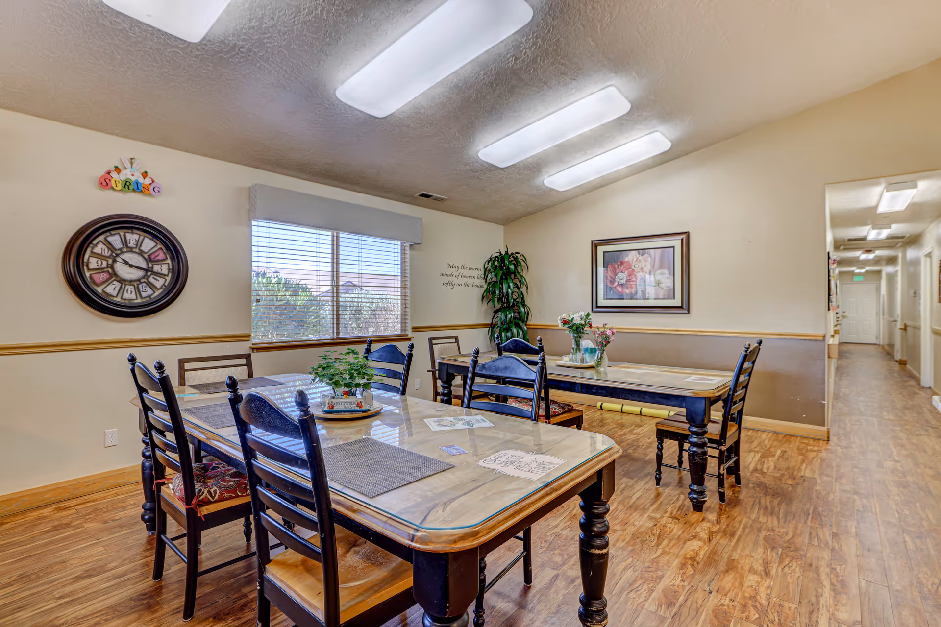 Bright dining room with multiple wooden tables and chairs, wall decor, a window, and overhead fluorescent lights.