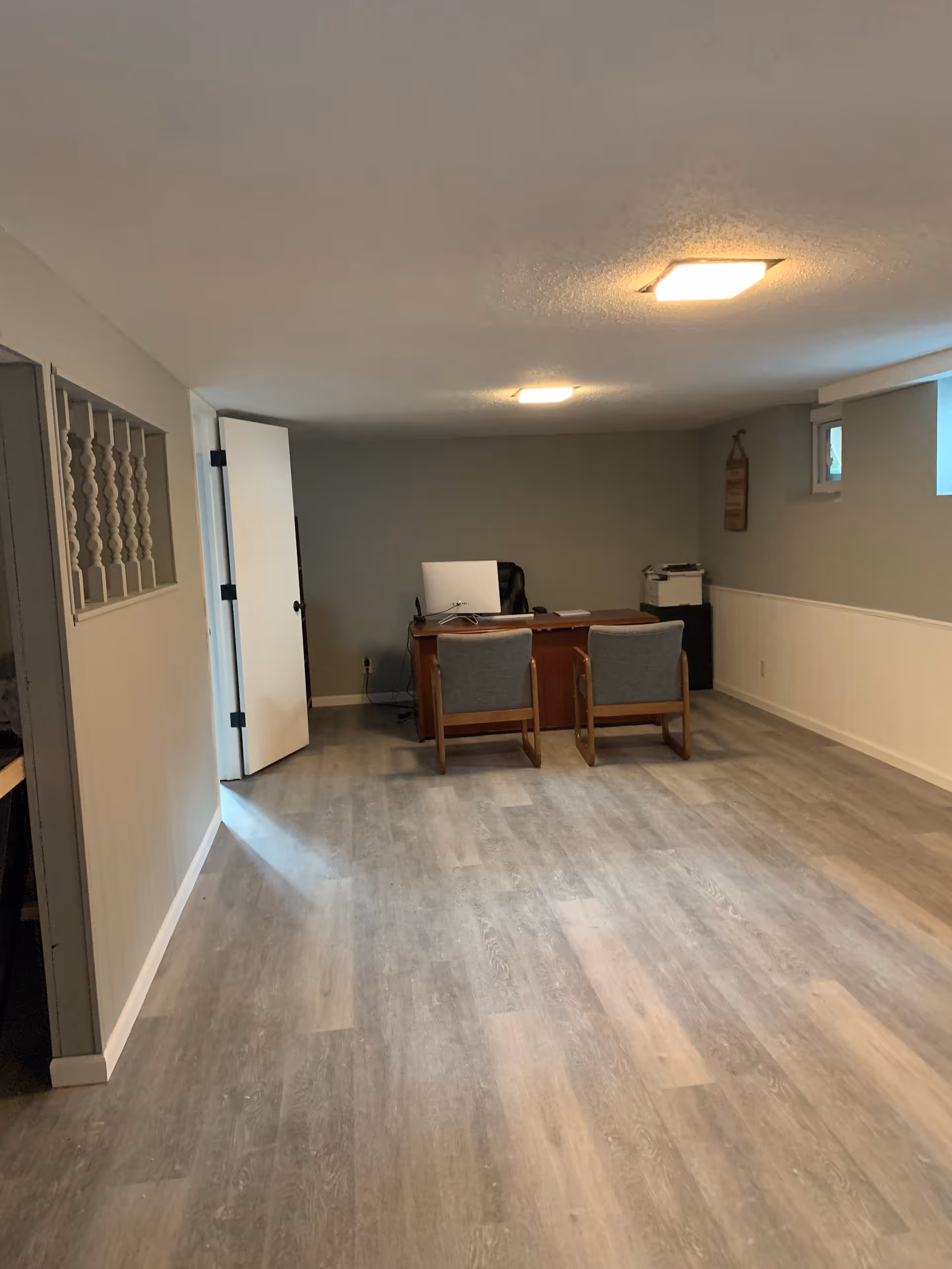 A small office room with a wooden desk, two gray cushioned chairs, a computer monitor, and a printer. The room has light gray walls, a white door slightly ajar, two small windows near the ceiling, and wood-patterned flooring.
