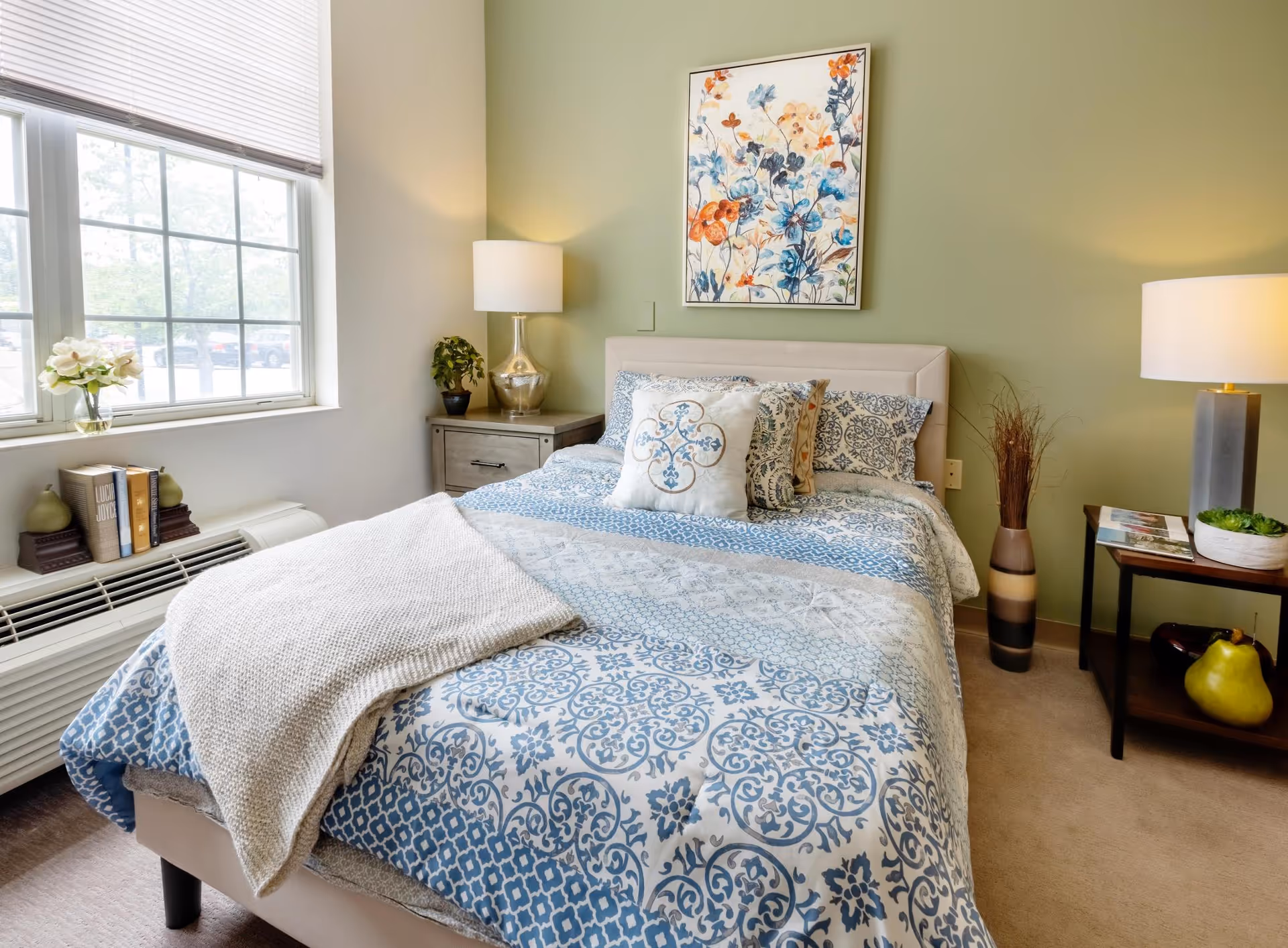 A cozy bedroom with a bed covered in blue and white patterned bedding and a beige throw blanket. There are two bedside tables with lamps, a small plant, books, and decorative items. A large window with blinds lets in natural light, and a colorful floral painting hangs on the green wall above the bed.