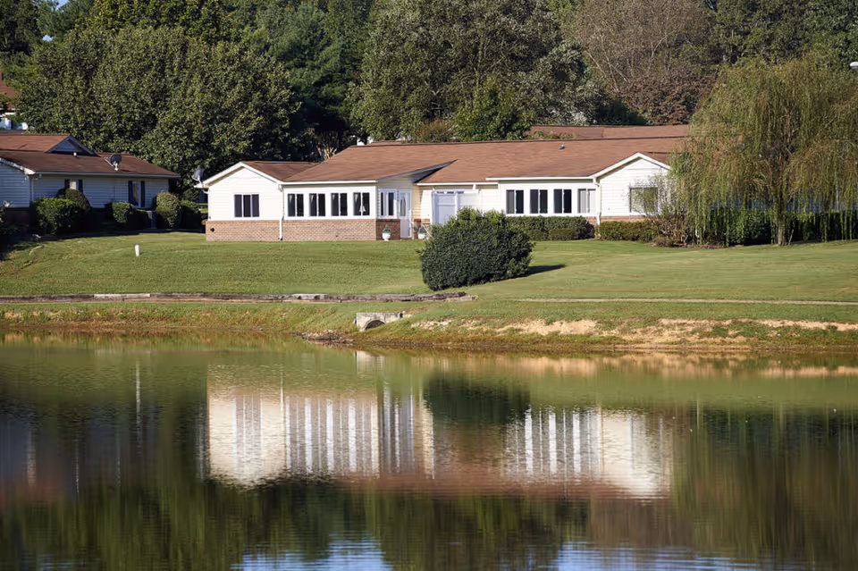 A single-story building with white siding and a brown roof situated on a grassy area near a calm pond, with trees in the background and the building's reflection visible in the water.