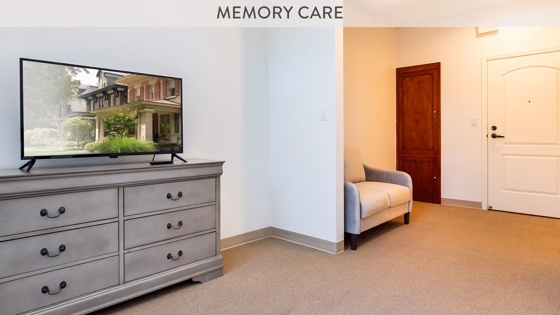 A memory care room with a gray dresser holding a flat-screen TV displaying a house exterior. There is a beige armchair next to a wooden cabinet and a white door in the background. The room has beige carpet and white walls.
